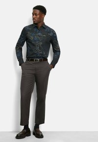 Langärmliges, schwarzes Shirt mit aufwendigem blauen Blumenmuster, kombiniert mit grauen Hosen und dunkelbraunen Schuhen. Glattes Material, taillierter Schnitt.