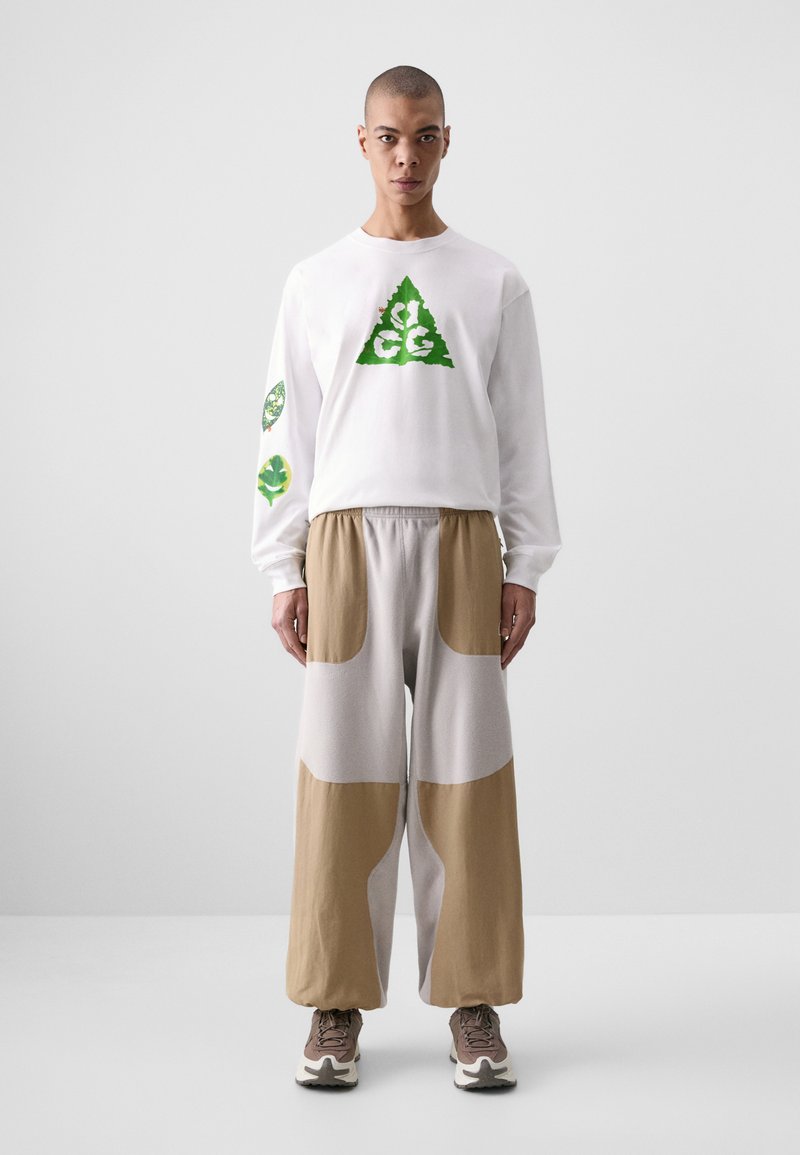 White long-sleeve shirt with green graphic designs, paired with loose grey and brown color-blocked pants and neutral sneakers.