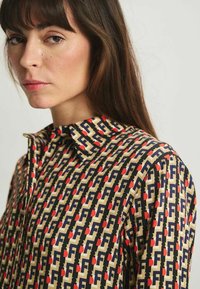 Blouse à motifs avec un fond beige et des motifs géométriques navy et rouge. Le col est de style classique, et le tissu semble lisse.