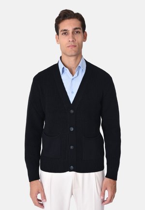 Cardigan nero lavorato a maglia con scollo a V, chiusura a sei bottoni, due tasche anteriori e polsini e orlo a coste. Indossato sopra una camicia azzurro chiaro.