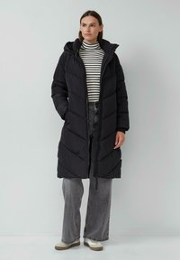 Zwarte pufferjas met een chevronontwerp, voorzien van een capuchon, een rits aan de voorkant en zijzakken, gedragen over een gestreepte coltrui en grijze broek.