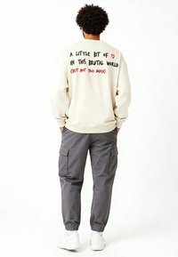 Felpa oversized beige con testo stampato sulla schiena, abbinata a pantaloni cargo grigi e sneakers bianche. Il testo presenta accenti rossi e neri.