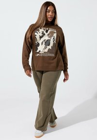 Brązowa bluza z grafiką w wzór skóry bydlęcej, zestawiona z oliwkowymi spodniami o szerokich nogawkach i jasnymi trampkami.