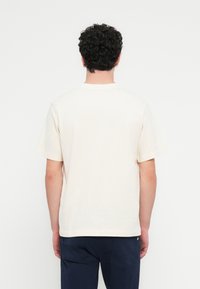 Μπεζ βαμβακερό μπλουζάκι με κλασικό λαιμόκο ψη, κοντά μανίκια και ίσια τελείωμα. Λεία υφή, χωρίς ορατά σχέδια ή γραφικά.