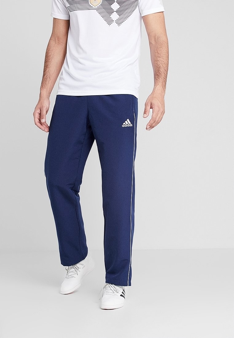 Man som bär marinblå Adidas träningsbyxor med vita ränder, tillsammans med vita sneakers och en vit t-shirt med grafiskt motiv.
