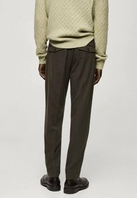 Un pantalon vert olive foncé avec une coupe ajustée, doté de poches arrière avec de discrètes fermetures à bouton, associé à un pull léger texturé.