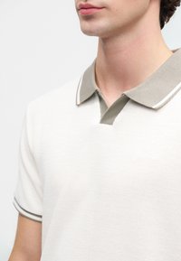 Junge Person in einem weißen Strickpolohemd mit grauem Kragen und Streifen an den Ärmeln, das bis zum Hals und oberen Brustbereich verkürzt ist.