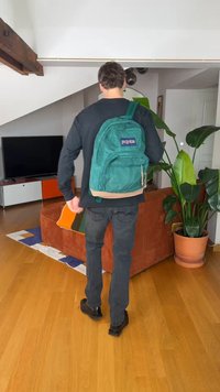 Osoba odchádzajúca v interiéri, nesúca tyrkysový batoh, oblečená v čiernom tričku a tmavých džínsach, blízko hnedej pohovky a veľkej kvetináča na drevenej podlahe.