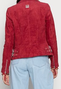 Červená sukně z semišové kůže se přiléhavým střihem, ozdobená stříbrnými detaily, bočními zipy a dvěma sponami v oblasti pasu.