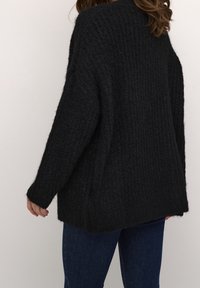 Musta, ylisuuri neulottu neule, jossa on tekstuurikuosi. Pitkät hihat ja leveä istuvuus, pidempi takasauma.