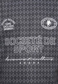 Mørk teksturert stoff med et diamantmønster i grå og svarte nyanser. Hvite bokstaver med "SOCIÉTÉ DE SPORT" og en sirkulær logodetalj.