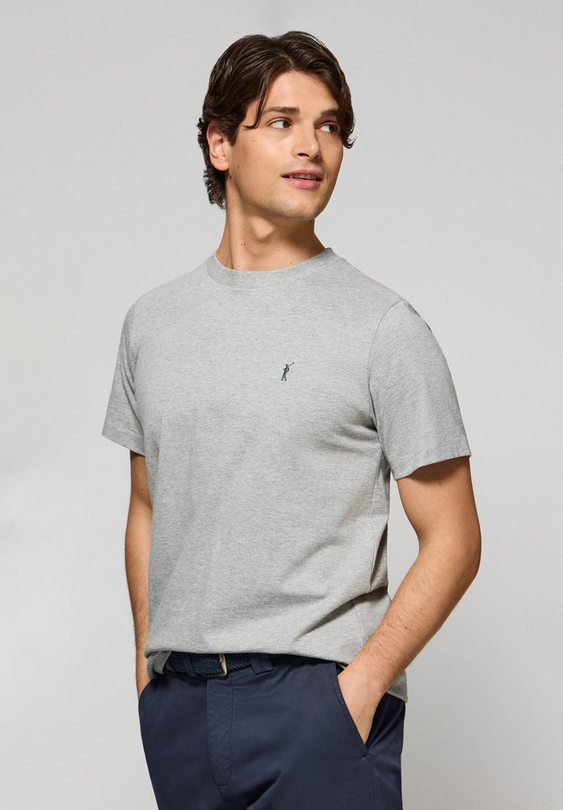 Camiseta de algodón gris con mangas cortas, cuello redondo, que presenta un pequeño logo bordado en azul marino. Textura suave y ajuste informal.