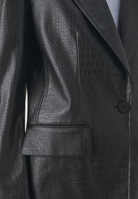 Blazer en similicuir noir avec texture crocodile, grands revers, fermeture par un bouton unique et détail d'une poche avant.