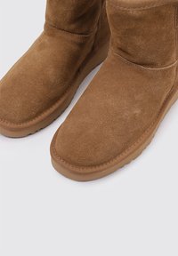 Botines de gamuza marrón con punta redondeada, textura suave y suela de goma plana. Presentan un diseño sencillo y sin costuras, con costuras visibles.