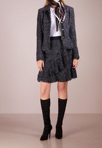 Meriino tweed takki, jossa on rispaantuneet yksityiskohdat, yhdistettynä vastaavaan röyhelömäiseen hameeseen. Mustat polvimittaiset ribatut saappaat ja raidallinen kaulahuivi korostavat asua.