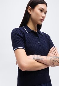 Νέα γυναίκα με μακριά σκούρα μαλλιά στέκεται με σταυρωμένα χέρια, φορώντας μπλε κοντομάνικο πόλο με λευκά περιθώρια και εμφανή τατουάζ στα αντιβράχια.