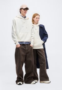 Νεαρός άνδρας και γυναίκα που φορούν κρεμ φούτερ με κουκούλα, καφέ ευρύχωρο παντελόνι, αθλητικά παπούτσια· η γυναίκα φορά επίσης ναυτικό παλτό τρεντς· και οι δύο στέκονται με τα χέρια στις τσέπες.