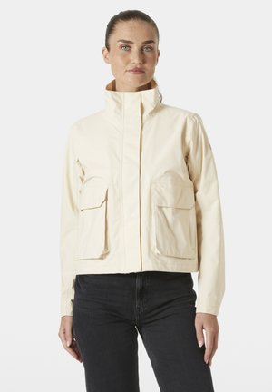 Vrouw met lichte, sproeterige huid en blauwe ogen, gekleed in een crèmekleurige jas met twee voorzakken en zwarte broek, staand tegen een effen achtergrond.