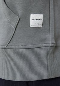 Sudadera gris con dobladillo y puños acanalados, con una etiqueta tejida que dice "JACK & JONES" cosida en el frente. Textura suave y lisa.