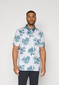 Blomster polo skjorte i hvit med tealkfin mønster, korte ermer og en ensfarget tealkrage. Laget av et lett og pustende stoff.