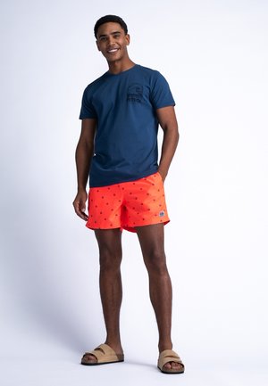 Marineblaues T-Shirt mit einem Logo; leuchtend orangefarbene Bade Shorts mit einem gepunkteten Muster; sandfarbene Slipper; Model steht vor einem schlichten Hintergrund.