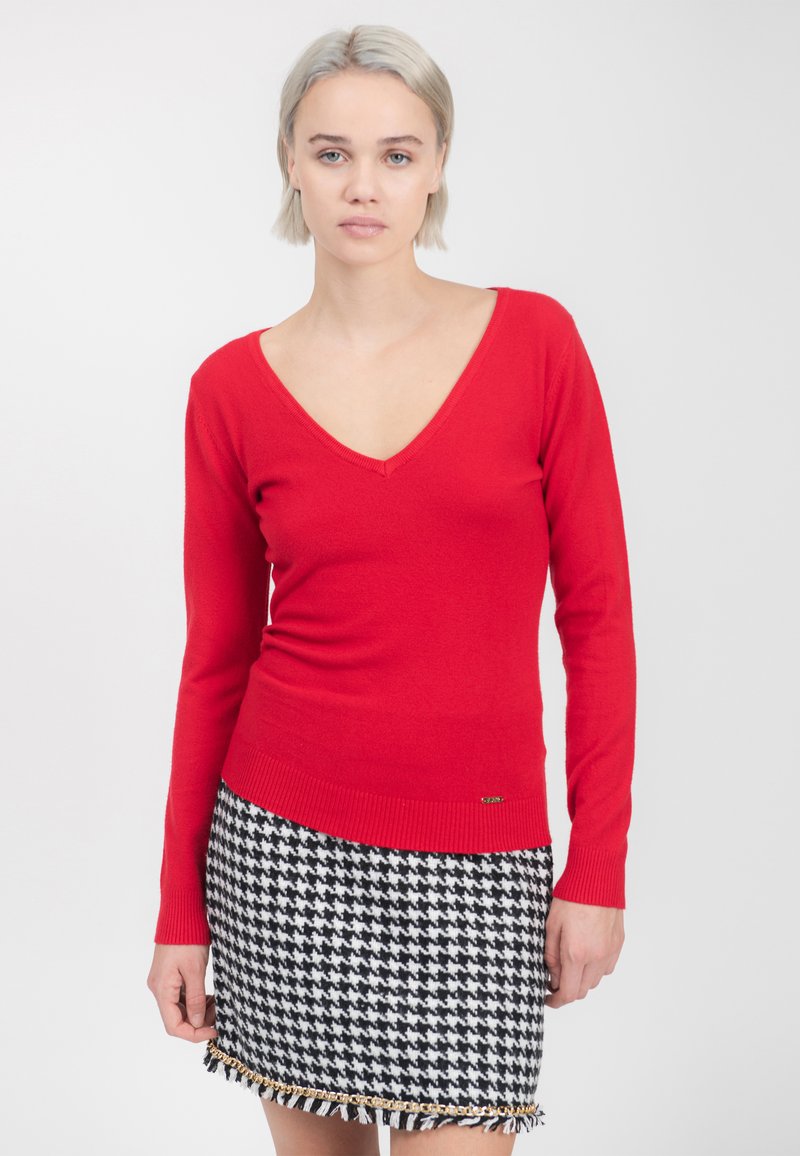 Roter V-Ausschnitt-Pullover, tailliert, mit langen Ärmeln. Kombiniert mit einem schwarz-weißen Hahnentrittmuster-Rock mit metallischen Akzenten und Fransen-Details.