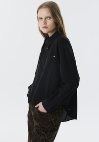Camicia nera oversize con bottoni, maniche lunghe, una tasca e dettagli in argento discreti, abbinata a pantaloni a stampa leopardata marroni.