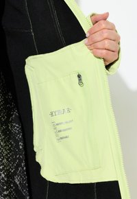 Veste vert clair, résistant à l'eau, avec une poche zippée. Les caractéristiques comprennent des étiquettes pour les propriétés imperméables, résistantes au vent et respirantes.