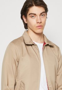 Chaqueta ligera de color marrón claro con cuello y cremallera frontal. Presenta un forro interior de cuadros y bolsillos laterales. Textura suave.