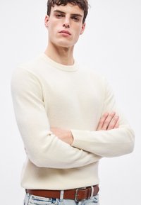 Krēmkrāsas džemperis ar apaļu izgriezumu, ribām aprocēs un apakšmalā. Kombinēts ar gaiši zilām džinsa biksēm un brūnu jostu ar metāla sprādzi.