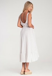 Gestreiftes, rückenfreies Maxikleid mit taillierter Taille und Rüschensaum. Hergestellt aus leichten Stoffen in sanften neutralen Tönen. Kombiniert mit Plateausandalen.