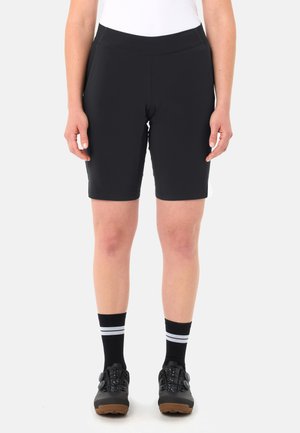 Persona che indossa pantaloncini neri aderenti fino al ginocchio, calzini neri con strisce bianche e scarpe sportive nere, in piedi su sfondo bianco.