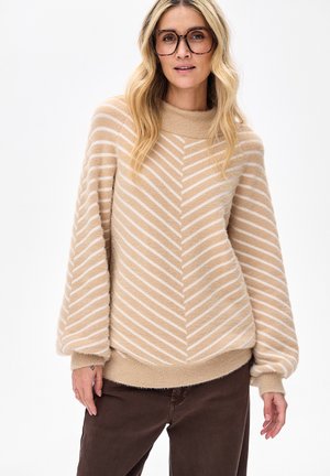 Blonde vrouw met een bril met bruin montuur en een beige-wit gestreepte, zachte trui met ballonmouwen en donkerbruine broek.