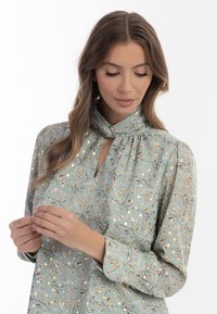 Blusa floreale in morbido tessuto verde chiaro con scollatura a chiave, maniche lunghe e delicati motivi pastello di fiori e uccelli.