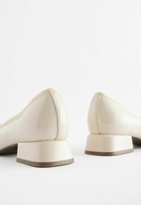 Chaussures en cuir verni beige avec un petit talon carré, bout rond et subtils accents texturés le long du bord. Fini lisse et brillant.