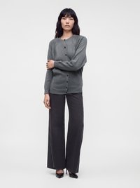 Жена, носеща сив кардиган с копчета, черен панталон с широки крачоли и страничен шев и черни обувки с остър връх, застанала на обикновен фон.