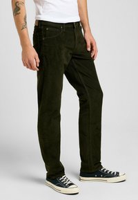 Pantaloni in velluto a coste verde scuro con gamba dritta, cinque tasche e bottoni metallici, abbinati a sneakers nere alte.