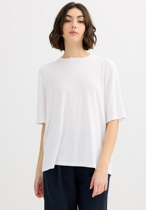 Mujer con cabello corto y castaño, vestida con una camiseta blanca lisa de manga corta y holgada y pantalones oscuros, de pie contra un fondo blanco.