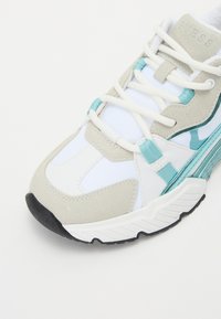 Fehér sneaker, textúrázott szürke velúr betéttel, aqua színű akcentusokkal és egy chunkyos fekete gumitalppal, amelynek zig-zag mintázata van.
