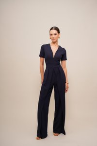 Marineblauwe jumpsuit met korte mouwen, diepe V-hals en geplooide voorkant. De stof lijkt glad; heeft brede, uitlopende pijpen en splitten.