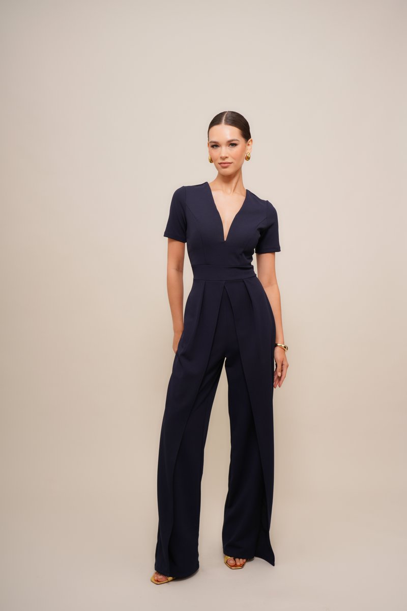 Marineblauwe jumpsuit met korte mouwen, diepe V-hals en geplooide voorkant. De stof lijkt glad; heeft brede, uitlopende pijpen en splitten.