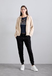 Béžová mikina na zips cez námornícku tričko so zlatým potlačou, doplnená čiernymi teplákmi a bielymi teniskami. Hladké textílie, uvoľnený strih.