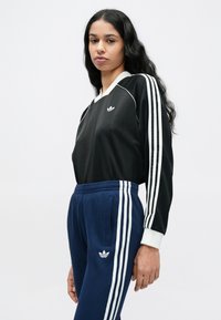 Fekete, hosszú ujjú felső fehér szegéllyel és Adidas logóval; hozzá illő sötétkék bordás melegítőnadrág fehér oldalsávval és logóval.