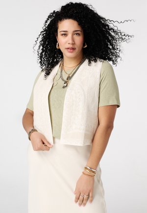 Femme aux cheveux bouclés portant une chemise vert clair, un gilet brodé crème, une jupe crème, des colliers superposés, des boucles d'oreilles créoles et des bracelets.