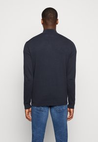 Uomo in piedi di spalle, che indossa un maglione scuro a maniche lunghe e jeans blu, su uno sfondo chiaro e uniforme.