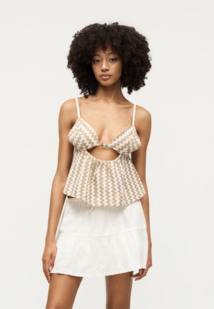 Νέα γυναίκα με σγουρά μαλλιά που φοράει μπεζ και λευκό crop top με ζιγκ-ζαγκ σχέδιο και μπροστινό άνοιγμα, καθώς και λευκή φούστα, στέκεται μπροστά σε απλό φόντο.