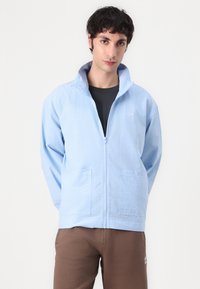 Light blue zip-up jacket made of textured fabric, featuring a high collar, two front pockets, and a small logo on the chest.