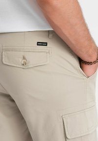 Shorts cargo beiges avec une poche à rabat et une fermeture à bouton, présentant une étiquette noire "ombre wear". Texture de tissu lisse et design structuré.