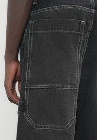 Μαύρο τζιν cargo παντελόνι με λευκή ραφή, διαθέτει μεγάλη πλαϊνή τσέπη και σχεδιασμό με στενό τελείωμα στο πόδι. Υφή υφάσματος με ομαλή επιφάνεια.