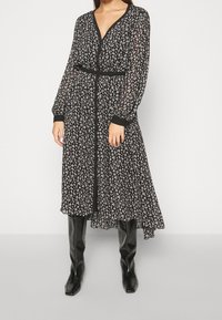 Musta kukallinen pitkä mekko pitkillä hihoilla, v-pääntiellä, kapealla vyötäröllä ja mustilla yksityiskohdilla. Yhdistetty polven yli ulottuviin mustiin saappaisiin.